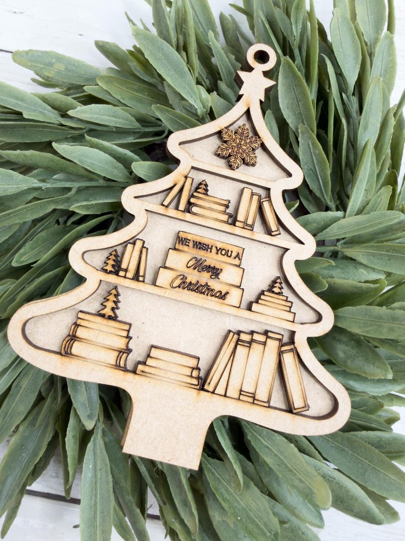 May include: A wooden Christmas tree ornament with a bookshelf design. The ornament features shelves filled with books and small Christmas trees. The text "WE WISH YOU A Merry Christmas" is visible. The ornament is light brown and has a snowflake at the top.
