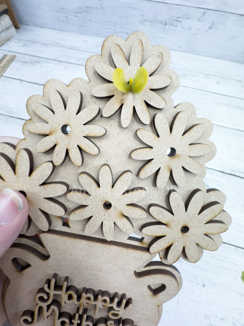May include: A wooden craft project featuring a vase with a bouquet of flowers. The vase is decorated with the words "Happy Mother's Day". A yellow butterfly is perched on top of the flowers.
