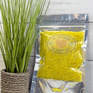 May include: A clear, sealed bag filled with yellow granules, labeled "DIY with Amber, The Makers Map." A potted green plant with long, thin leaves sits to the left. The bag is on a light-colored wooden surface.