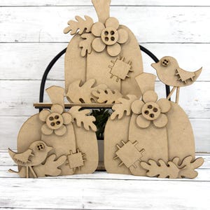 May include: Three unfinished wooden pumpkins with leaves, flowers, and buttons. A small bird with a button eye is perched on a branch.
