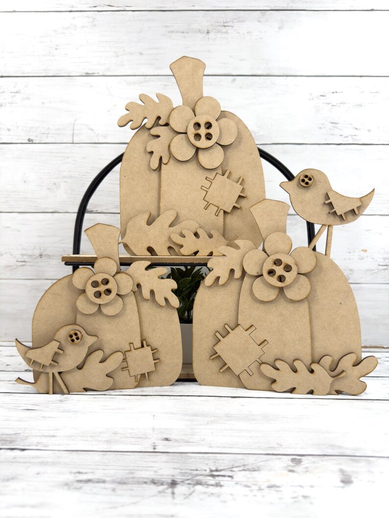 May include: Three unfinished wooden pumpkins with leaves, flowers, and buttons. A small bird with a button eye is perched on a branch.