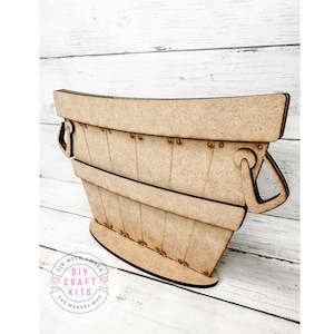 May include: A wooden DIY craft kit shaped like a bucket, featuring a curved top and base, with two handle cutouts. The bucket is constructed with vertical slats and horizontal bands, and is held together with small rivets. The kit includes the text "DIY WITH AMBER" and "DIY CRAFT KITS".