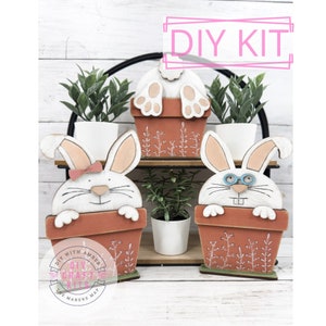 May include: DIY craft kit featuring three wooden bunny planter decorations. The bunnies are painted white with black eyes and noses. One bunny has a pink bow, another has blue glasses, and the third is peeking out of the planter from behind. The planters are painted brown with green plant designs.