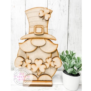 May include: A wooden leprechaun-themed craft kit. The unfinished wood figure features a tall hat with a shamrock, a beard, and shamrocks. The base includes the text "DIY WITH AMBER DIY CRAFT KITS THE MAKERS MAP".