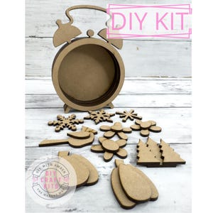 May include: DIY craft kit featuring a wooden alarm clock with a circular cutout and various wooden shapes, including snowflakes, flowers, and Christmas trees. The kit is labeled "DIY with Amber, DIY Craft Kits, The Makers Market".