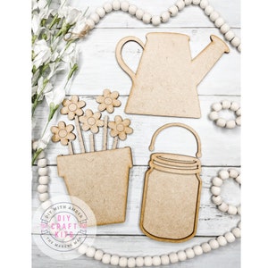 May include: Wooden craft kit featuring a watering can, a mason jar, and a flower pot with flowers. The kit is unfinished and ready to be painted or stained. DIY with Amber, The Makers Map, DIY Craft Kits.