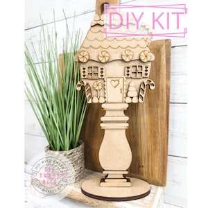 May include: A wooden gingerbread house craft kit with candy cane details, a heart-shaped door, and a candy lollipop. The house is on a wooden stand with a base.