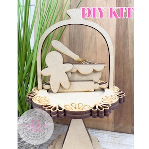 May include: A wooden DIY kit featuring a gingerbread man, mixing bowl, rolling pin, and whisk. The kit includes a decorative base with a floral design and a handle. The words "DIY KIT" are displayed at the top.