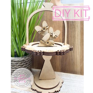 May include: A DIY kit for a wooden cake stand with a decorative heart design. The stand features a round base, a pedestal, and a scalloped edge. The top tier has heart-shaped cutouts and is adorned with buttons. The words "DIY KIT" are visible.
