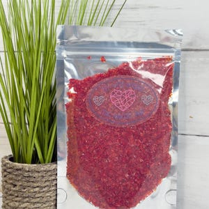 May include: A clear, sealed bag filled with red, granular material. A clear label with a pink heart design and text is affixed to the bag. A potted green plant is to the left of the bag, against a white wood-paneled background.