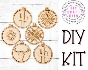 May include: Six unfinished wooden ornaments with various designs, including a cactus, a snowflake, a geometric pattern, and a bull's head. The ornaments are arranged on a white wooden surface. The text "DIY CRAFT KITS" is visible in a circle with a pink background. The text "DIY KIT" is also visible in large, black letters with a pink outline.