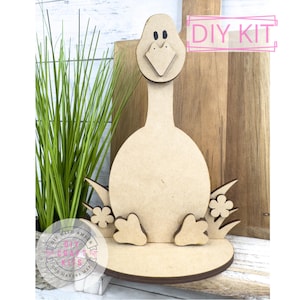 May include: DIY craft kit featuring a wooden cutout of a goose with a beak, eyes, and feet. The goose is sitting on a round base with grass and flowers around it. The kit includes all the pieces needed to assemble the goose.