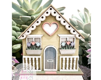 May include: A wooden house shaped planter with a heart cutout on the front, two windows with flower boxes, and a door. The planter is decorated with white trim and pink accents. The planter is sitting on a bed of white sand.
