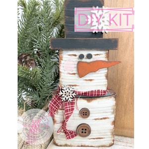 May include: A handcrafted snowman DIY kit made from repurposed materials. The snowman features a black top hat, an orange carrot nose, and a red and white plaid scarf. The text "DIY KIT" is visible on the top. Two large wooden buttons serve as the snowman's buttons.