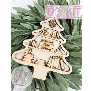 May include: A wooden Christmas tree-shaped DIY kit with shelves of books and the text "We Wish You a Merry Christmas." The kit includes a snowflake and small Christmas trees. The background features green foliage, and the kit is labeled "DIY KIT."