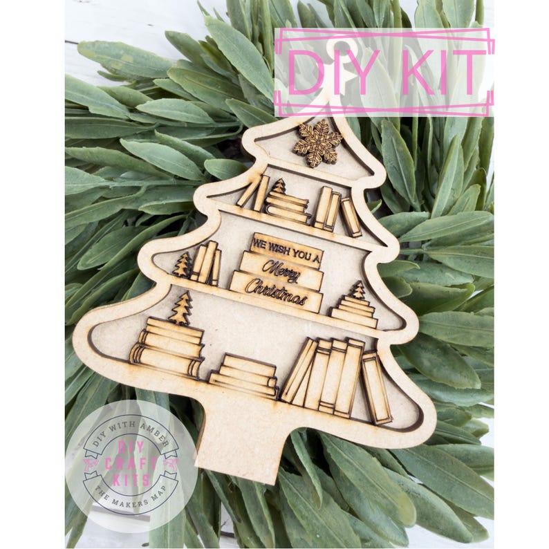 May include: A wooden Christmas tree-shaped DIY kit with shelves of books and the text "We Wish You a Merry Christmas." The kit includes a snowflake and small Christmas trees. The background features green foliage, and the kit is labeled "DIY KIT."