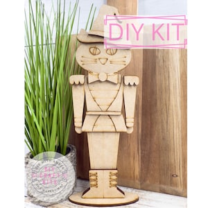 May include: A wooden DIY kit featuring a cat-shaped nutcracker wearing a hat and bow tie. The cat has cut-out details for eyes, nose, and whiskers. The words "DIY KIT" are in a pink box. The kit is displayed with a plant in a pot.
