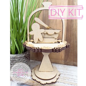 May include: A wooden DIY kit featuring a tiered cake stand with gingerbread man, rolling pin, whisk, and mixing bowl cutouts. The stand has a decorative scalloped edge and the text "DIY KIT".