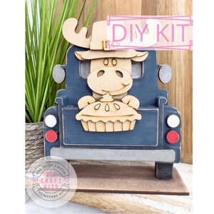 May include: A wooden DIY kit featuring a blue truck with a smiling moose holding a pie. The moose has antlers and is wearing a hat. The kit includes the text "DIY KIT" in a pink rectangle. The base is brown, and the wheels have red and white accents.