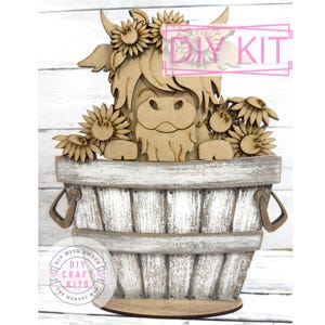 May include: A wooden DIY kit featuring a Highland cow with sunflowers, set in a rustic bucket. The cow is surrounded by laser-cut sunflowers and the text "DIY KIT" is visible. The bucket has metal handles and a distressed finish.