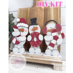 May include: DIY kit featuring three wooden snowman figures. Each snowman has a carrot nose, black dot eyes, and holds a festive item: a candy cane, a donut, and earmuffs. The text "DIY KIT" is at the top.