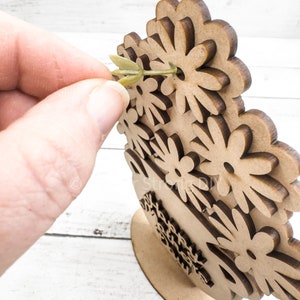 May include: A wooden craft project featuring a vase with a floral design. The vase is decorated with multiple flower shapes and has a base for standing. A green stem is being inserted into one of the flower shapes.