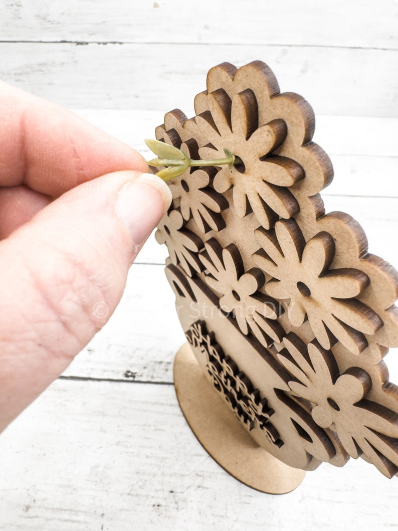 May include: A wooden craft project featuring a vase with a floral design. The vase is decorated with multiple flower shapes and has a base for standing. A green stem is being inserted into one of the flower shapes.