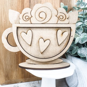 Op de afbeelding: Houten uitsnijding van een theekopje met een handvat en drie harten erin. Het theekopje is versierd met een swirl-ontwerp en heeft een aparte houten basis.