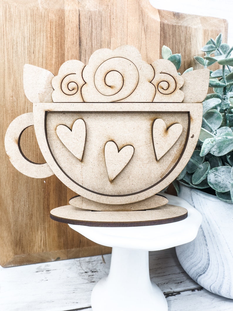 Op de afbeelding: Houten uitsnijding van een theekopje met een handvat en drie harten erin. Het theekopje is versierd met een swirl-ontwerp en heeft een aparte houten basis.
