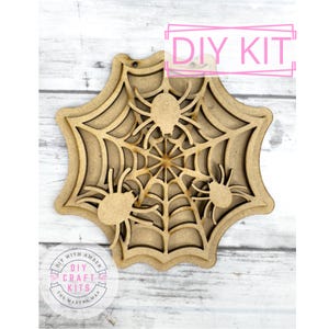 May include: A wooden DIY kit featuring a layered spider web design with three spiders. The words "DIY KIT" are in a pink box at the top. The kit is designed for crafting and is perfect for Halloween decorations.
