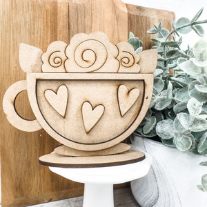 Op de afbeelding: Houten uitsnijding van een koffiekopje met een swirl-ontwerp aan de bovenkant en drie harten aan de voorkant. Het kopje staat op een wit voetstuk.