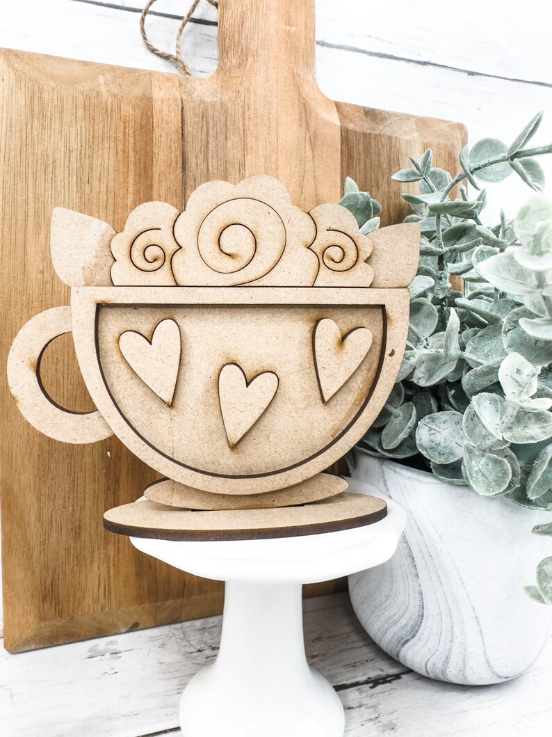 Op de afbeelding: Houten uitsnijding van een koffiekopje met een swirl-ontwerp aan de bovenkant en drie harten aan de voorkant. Het kopje staat op een wit voetstuk.