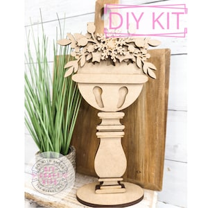 May include: DIY craft kit featuring a wooden cutout of a vase with a floral arrangement. The vase is on a pedestal base and the flowers are detailed with snowflakes and berries. The kit is labeled "DIY Kit".