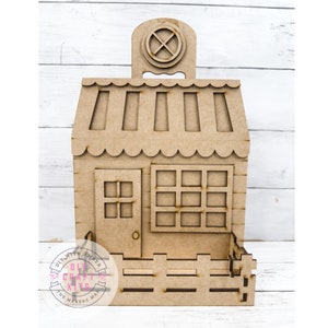 May include: A wooden craft kit shaped like a small house with a sign above the door that says "DIY with Amber Craft Kits The Makers Map". The house has a door, windows, and a fence around the bottom.