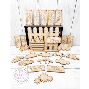 May include: A set of wooden craft blanks with various phrases and designs, including "Happy Holidays", "Winter Wishes", "April Showers", "Made in America", "Love You Lots", and "Back to School". The blanks are arranged on a wooden fence with a small wooden tree and a heart shape.