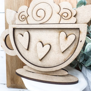Op de afbeelding: Houten uitsnijding van een koffiekopje met een swirl-ontwerp aan de bovenkant en drie harten erin. Het kopje staat op een houten basis.