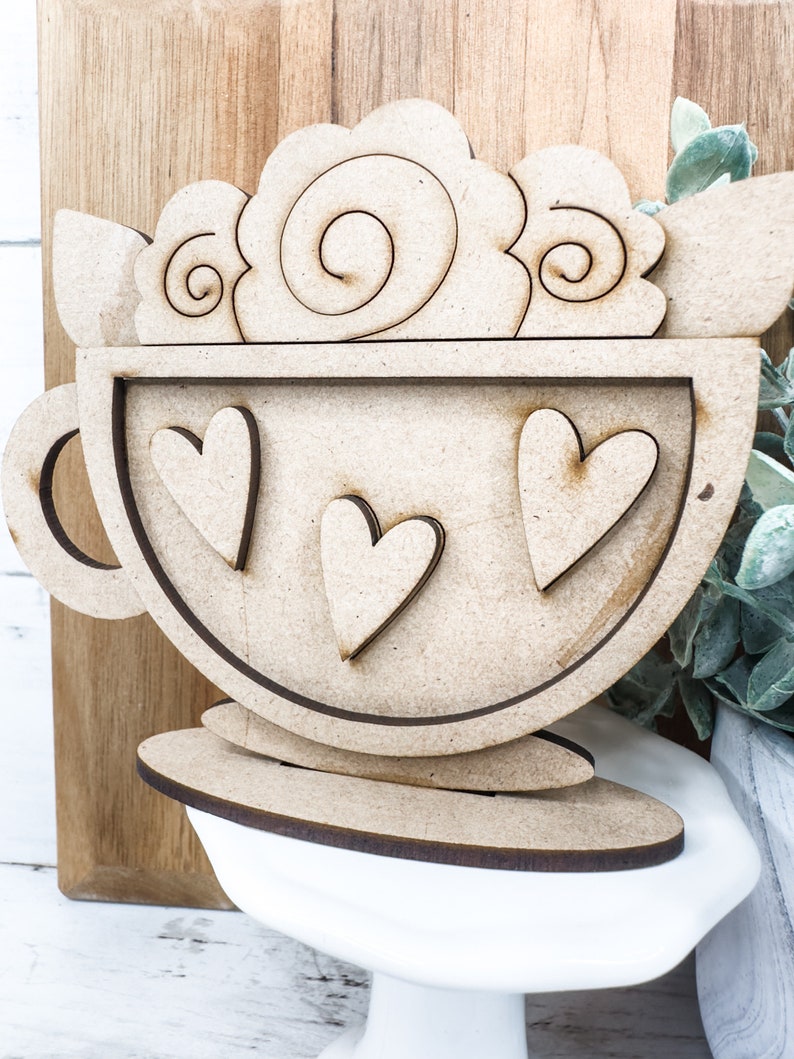 Op de afbeelding: Houten uitsnijding van een koffiekopje met een swirl-ontwerp aan de bovenkant en drie harten erin. Het kopje staat op een houten basis.