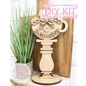 May include: A wooden DIY craft kit to create a Christmas decoration. The kit includes a wooden cup with the words "Sweet Wishes" and a candy cane design, a wooden stand, and a wooden base. The kit is unfinished and ready to be painted or stained.