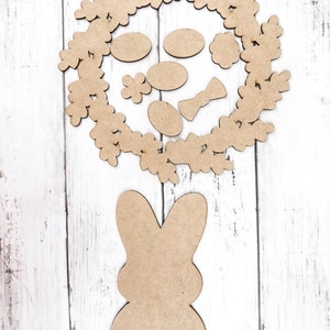 May include: A brown wooden bunny cutout with a wreath of leaves and flowers around it. The wreath has a bow and several oval shapes inside.