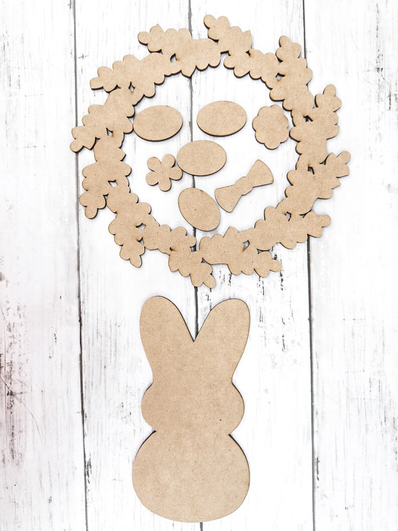 May include: A brown wooden bunny cutout with a wreath of leaves and flowers around it. The wreath has a bow and several oval shapes inside.