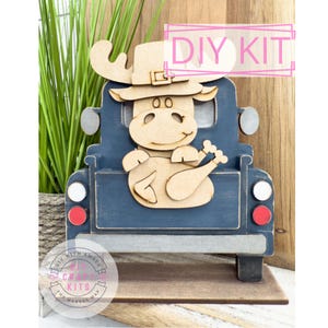 May include: A wooden DIY kit featuring a blue truck with a moose wearing a pilgrim hat and holding a turkey. The kit includes the text "DIY KIT" in a pink rectangle. The base has the text "DIY WITH AMBER" and "DIY CRAFT KITS THE MAKERS MAP".