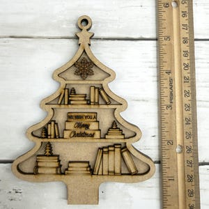 May include: A wooden Christmas tree ornament with a bookshelf design. The ornament features shelves filled with books and the text "WE WISH YOU A Merry Christmas." A ruler is next to the ornament, measuring in inches.