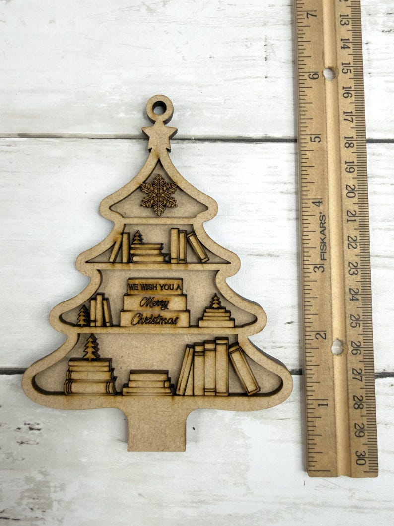 May include: A wooden Christmas tree ornament with a bookshelf design. The ornament features shelves filled with books and the text "WE WISH YOU A Merry Christmas." A ruler is next to the ornament, measuring in inches.