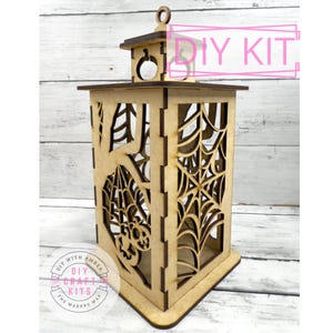 May include: A wooden DIY kit lantern with a Halloween theme. The lantern features cut-out designs of spiderwebs and a skull. The text "DIY KIT" is visible. The lantern is made of light brown wood and has a rectangular shape.