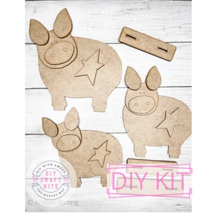 May include: DIY craft kit featuring three unfinished wooden pig shapes with stars. The kit includes a wooden strip for assembling the pigs. The kit is labeled "DIY Craft Kits The Makers Map" and "DIY Kit".