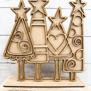 Op de afbeelding: Houten kerstboomuitsparing met een standaard. De bomen zijn versierd met swirls, harten en stippen. De bomen zijn bruin en hebben een rustieke uitstraling.