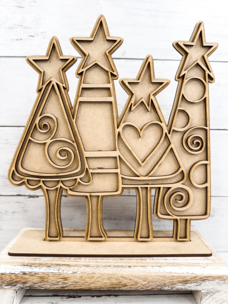 Op de afbeelding: Houten kerstboomuitsparing met een standaard. De bomen zijn versierd met swirls, harten en stippen. De bomen zijn bruin en hebben een rustieke uitstraling.