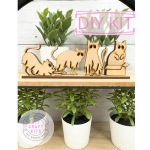May include: A DIY kit featuring wooden cat figurines in various poses, including one in a box, displayed on a shelf with potted plants. The text "DIY KIT" is visible. The kit is from "DIY with Amber Craft Kits".