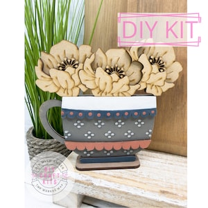 May include: DIY kit featuring a wooden teacup-shaped planter with laser-cut flowers. The teacup is painted gray with white and pink accents. The text "DIY KIT" is displayed in a pink rectangle.
