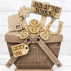 May include: Wooden craft decoration featuring a basket filled with gold coins, a shamrock, a leprechaun hat, a horseshoe, and a sign that reads "End of the Rainbow". The basket has a tag that says "Lucky You".
